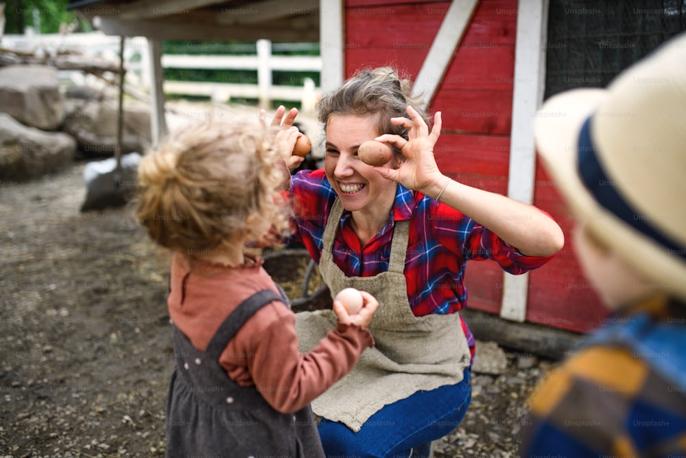 Madre alegre con niños pequeños de pie en la granja, divirtiéndose al recolectar huevos.