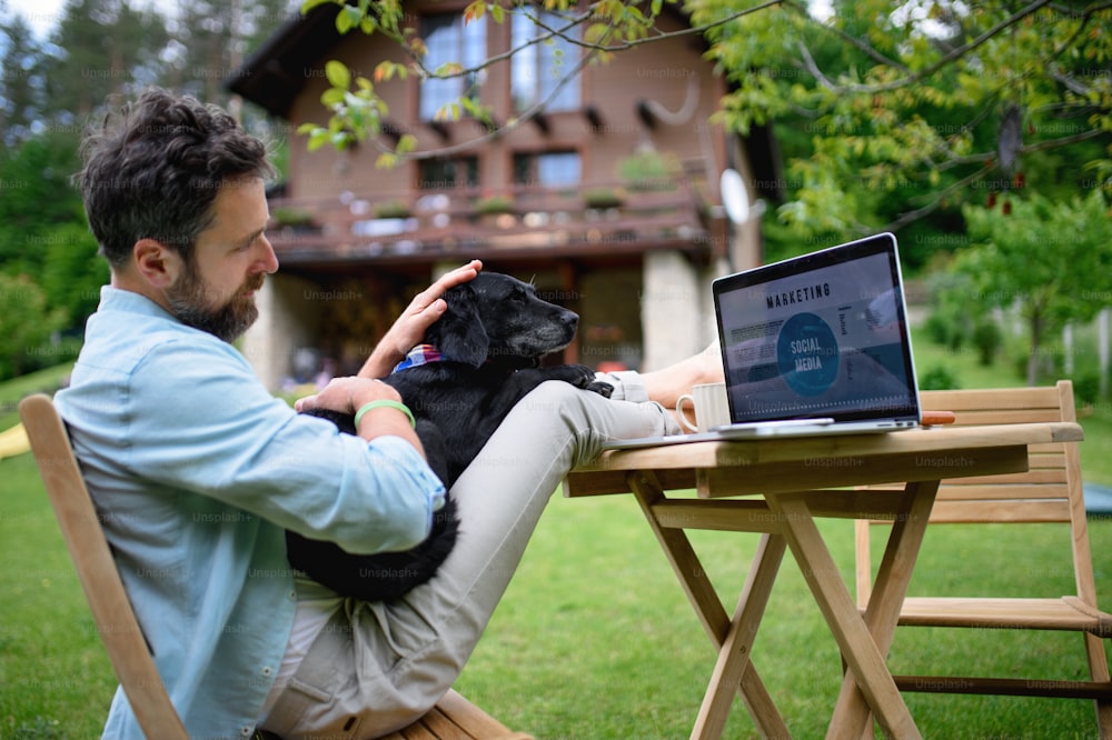 Happy mature man with laptop and dog working outdoors in garden, home office concept.