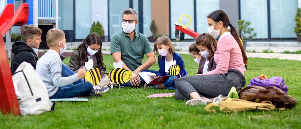 Gruppe fröhlicher Kinder mit Lehrer mit Gesichtsmasken, die nach der Covid-19-Quarantäne und -Sperrung im Freien in der Schule lernen.