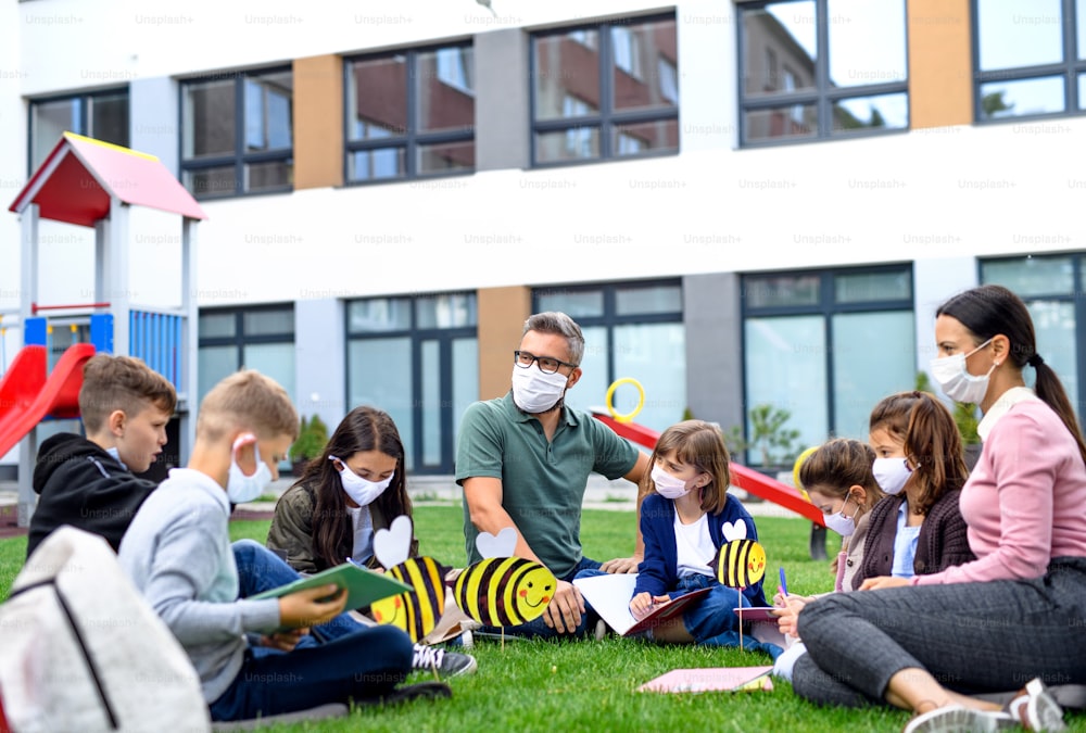 Grupo de niños alegres con maestro y máscaras faciales aprendiendo al aire libre en la escuela después de la cuarentena y el bloqueo de covid-19.