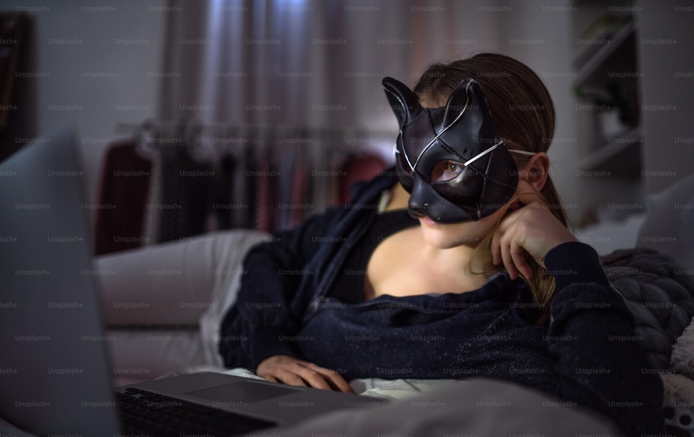 Vorderansicht eines jungen Mädchens mit Katzenmaske und Laptop in Innenräumen, Online-Dating und Missbrauchskonzept.