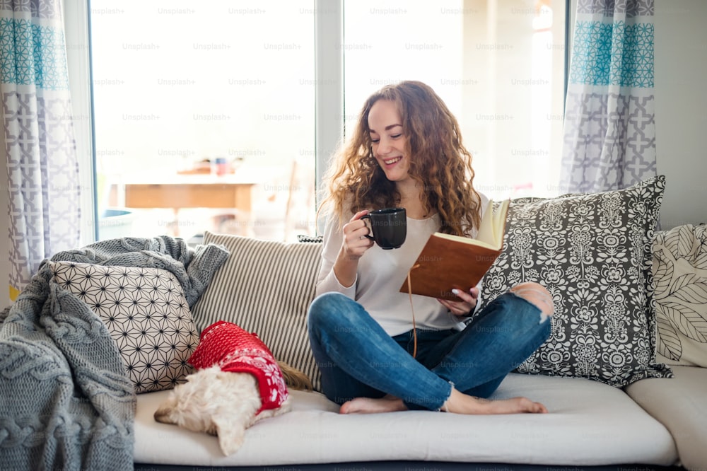 Jeune femme avec chien et livre relaxant sur canapé à l’intérieur à la maison, lecture.