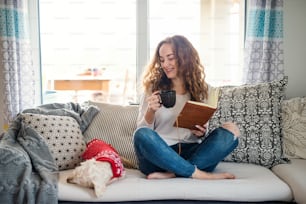 Mujer joven con perro y libro relajándose en el sofá en el interior de casa, leyendo.