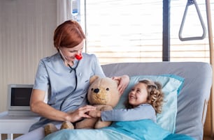 Amable doctora con estetoscopio que examina a una niña pequeña en la cama en el hospital.