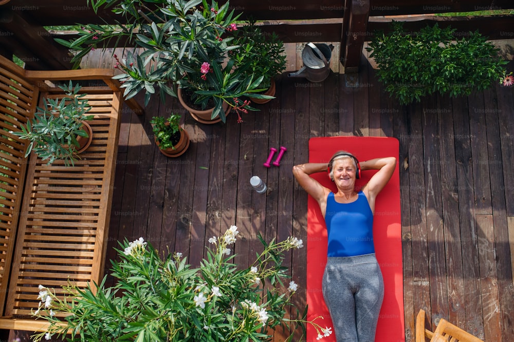 A top view of senior woman with headphones outdoors on a terrace in summer, resting after doing exercise on mat.