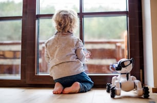 Eine Rückansicht eines niedlichen Kleinkindes, das zu Hause drinnen auf dem Boden sitzt und mit einem Roboterhund spielt.