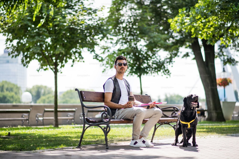 Junger blinder Mann mit weißem Stock und Blindenhund sitzt auf einer Bank im Park in der Stadt und ruht sich aus.