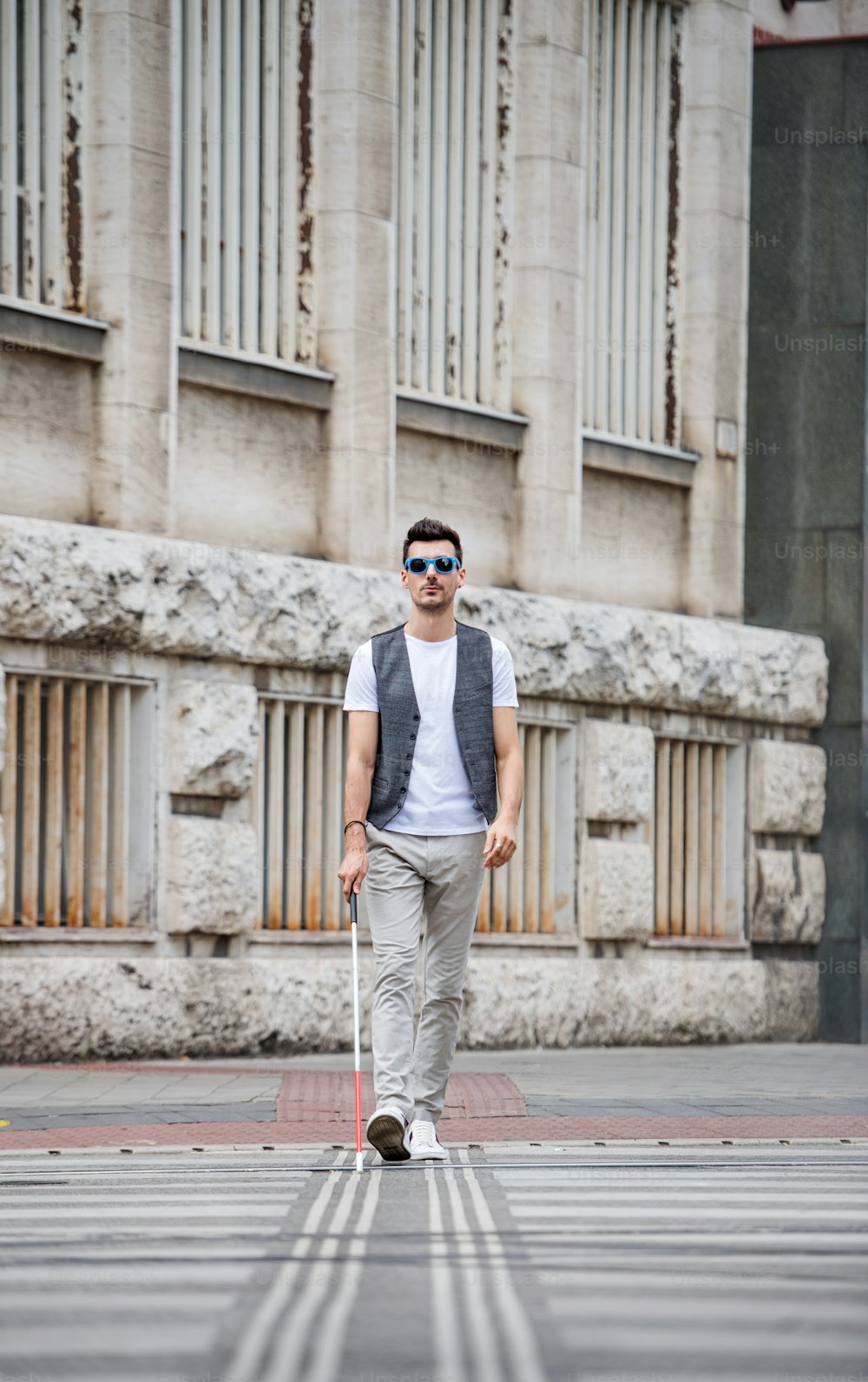 Young blind man with white cane walking across the street in city. Copy space.