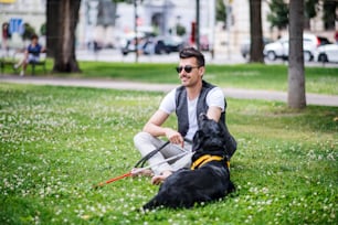 Young blind man with white cane and guide dog sitting in park in city, resting.