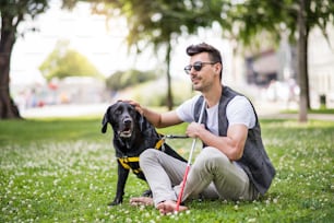Joven ciego con bastón blanco y perro guía sentado en el parque de la ciudad, descansando.