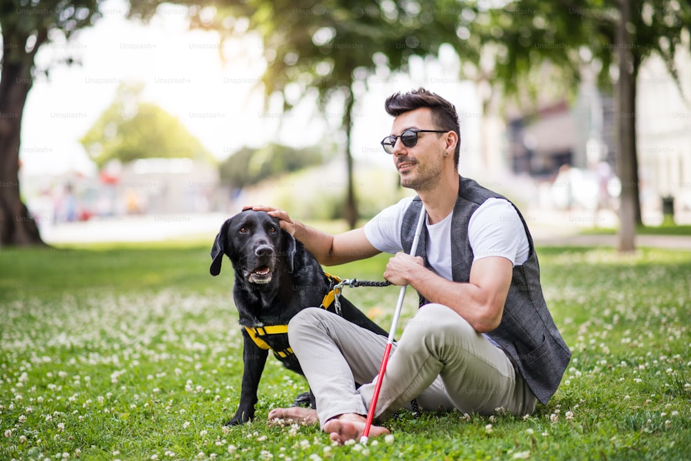 白い杖と盲導犬を持つ若い盲目の男性が街の公園に座って休んでいる。