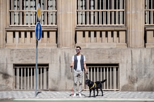Joven ciego con bastón blanco y perro guía parado en el pavimento de la ciudad, esperando.