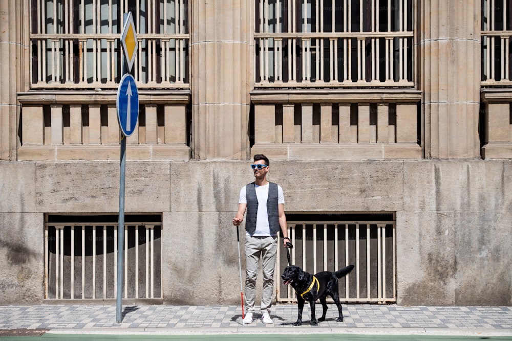 Young blind man with white cane and guide dog standing on pavement in city, waiting.