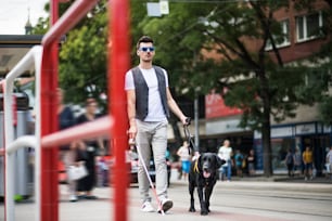 Ein junger blinder Mann mit weißem Stock und Blindenhund geht in der Stadt auf dem Bürgersteig spazieren.