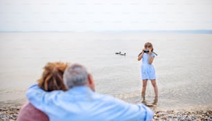 A small girl with camera and grandparents on holiday ba lake, taking photos.