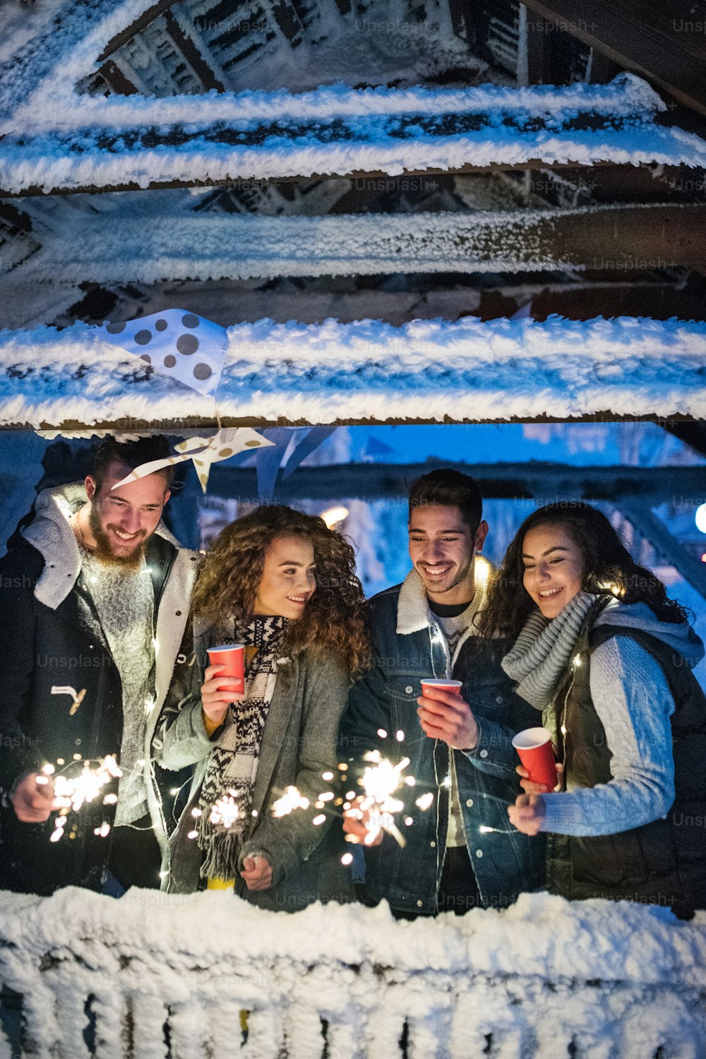 A group of young friends outdoors in snow in winter at night, holding sparklers.