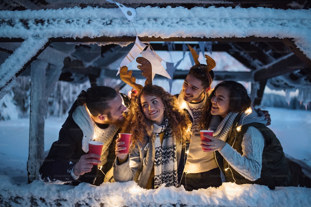 Un groupe de jeunes amis dehors dans la neige en hiver la nuit, tenant des boissons.