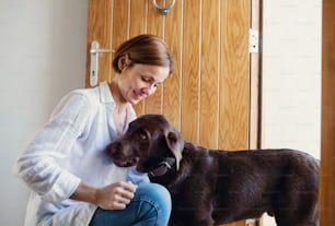Eine glückliche junge Frau, die zu Hause drinnen an der Tür auf dem Boden sitzt und mit einem Hund spielt.