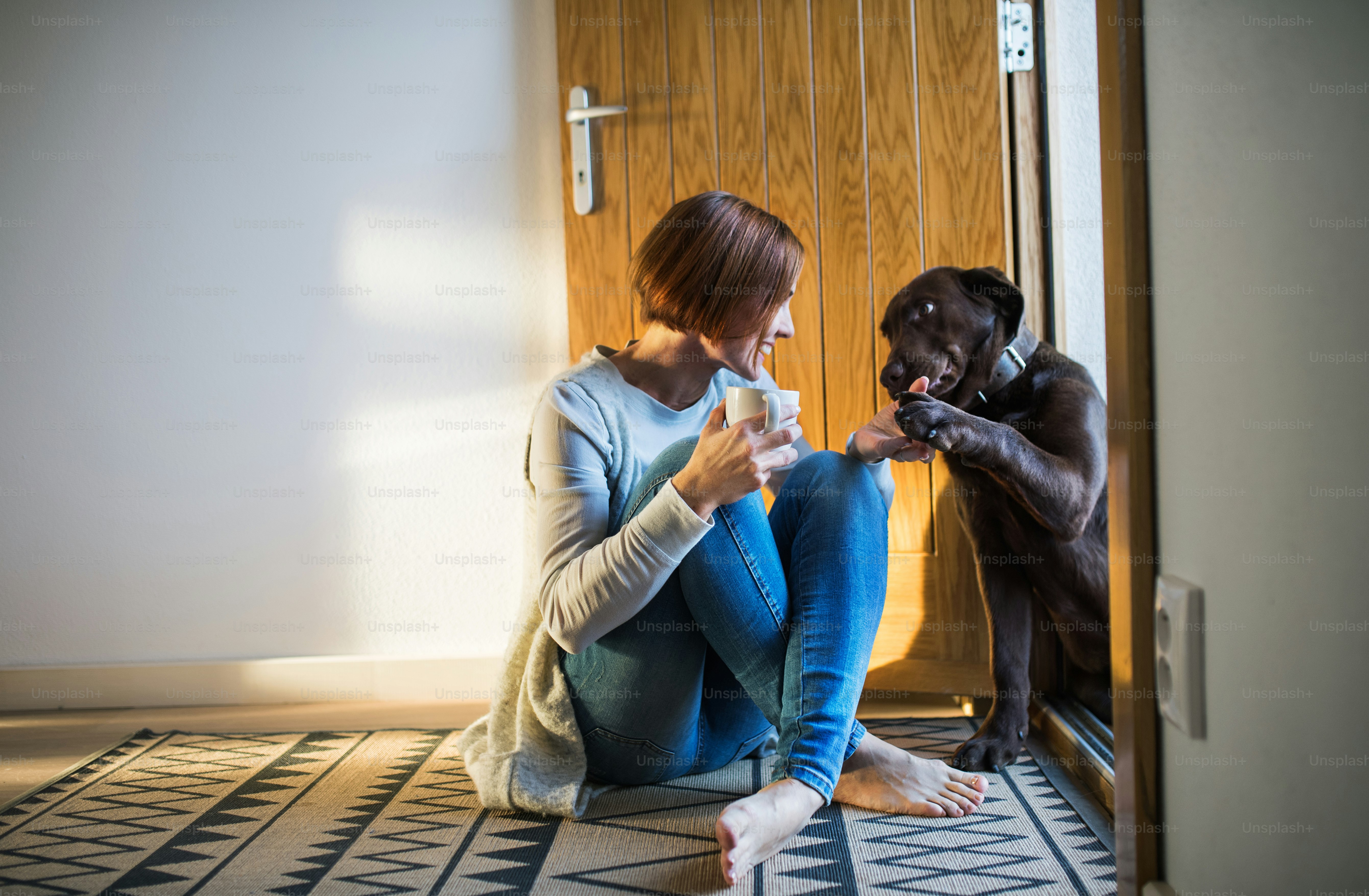 A young woman with a cup of coffee sitting indoors by the door on the floor at home, playing with a dog. Copy space