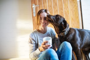 A young woman with a cup of coffee sitting indoors on the floor at home, playing with a dog.