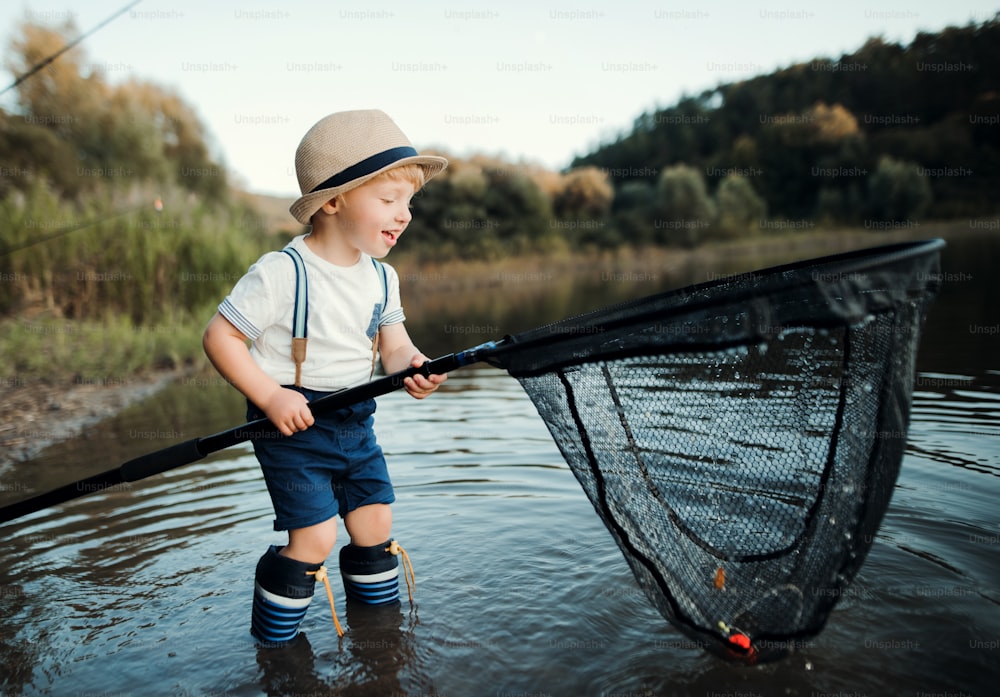 A happy small toddler boy standing in water and holding a net by a lake, fishing.