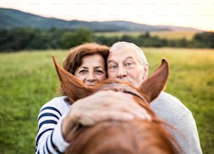 A crazy senior couple standing by a horse outside in nature, looking over his head, holding it.
