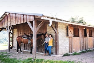 A senior couple with a brown horse standing in front of a wooden stable.