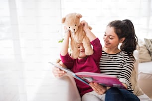 Giovane bella madre e una bambina con l'orsacchiotto a casa, leggendo un libro.