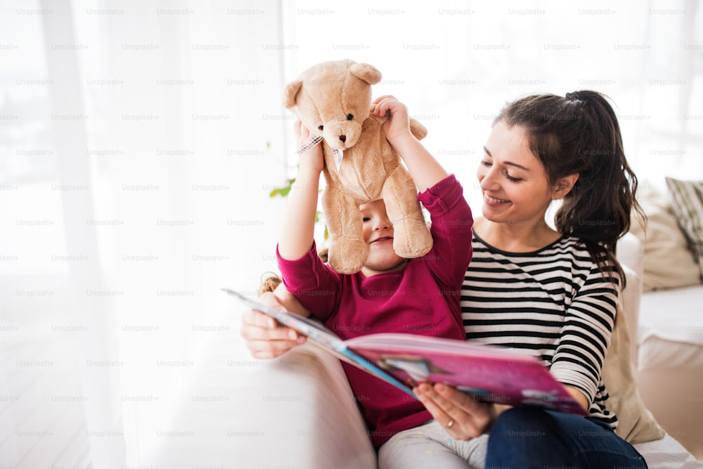 Jovem bela mãe e uma menina pequena com ursinho de pelúcia em casa, lendo um livro.