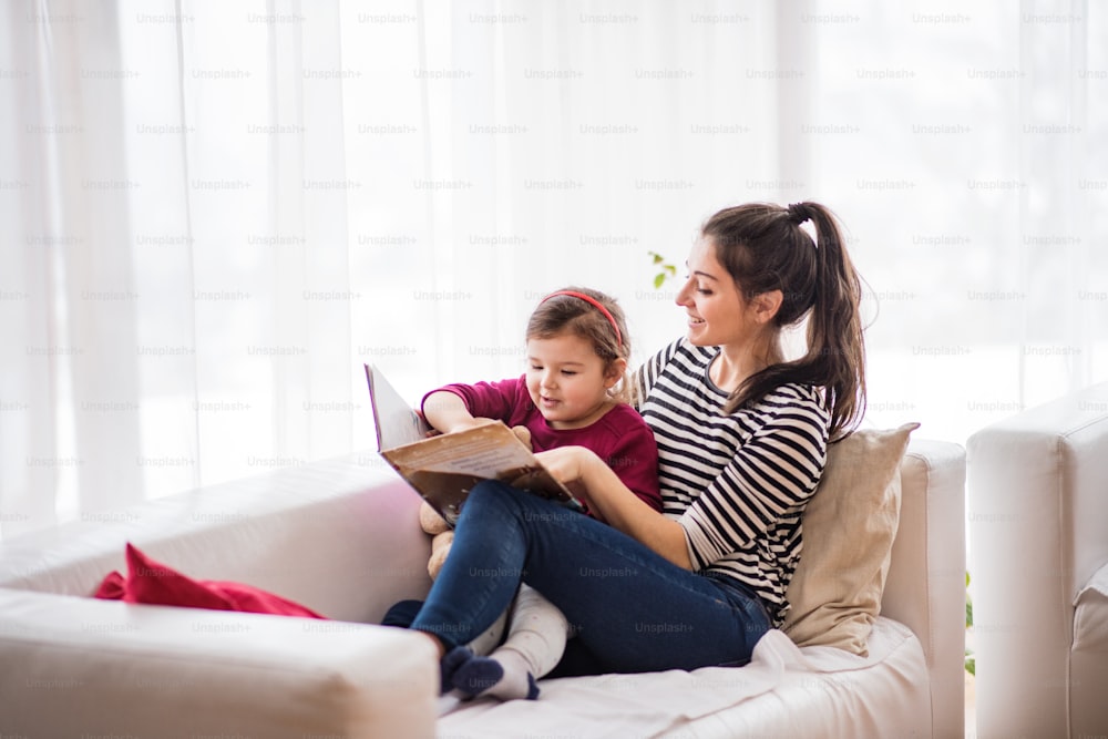 Joven y hermosa madre con una niña pequeña en casa, leyendo un libro.