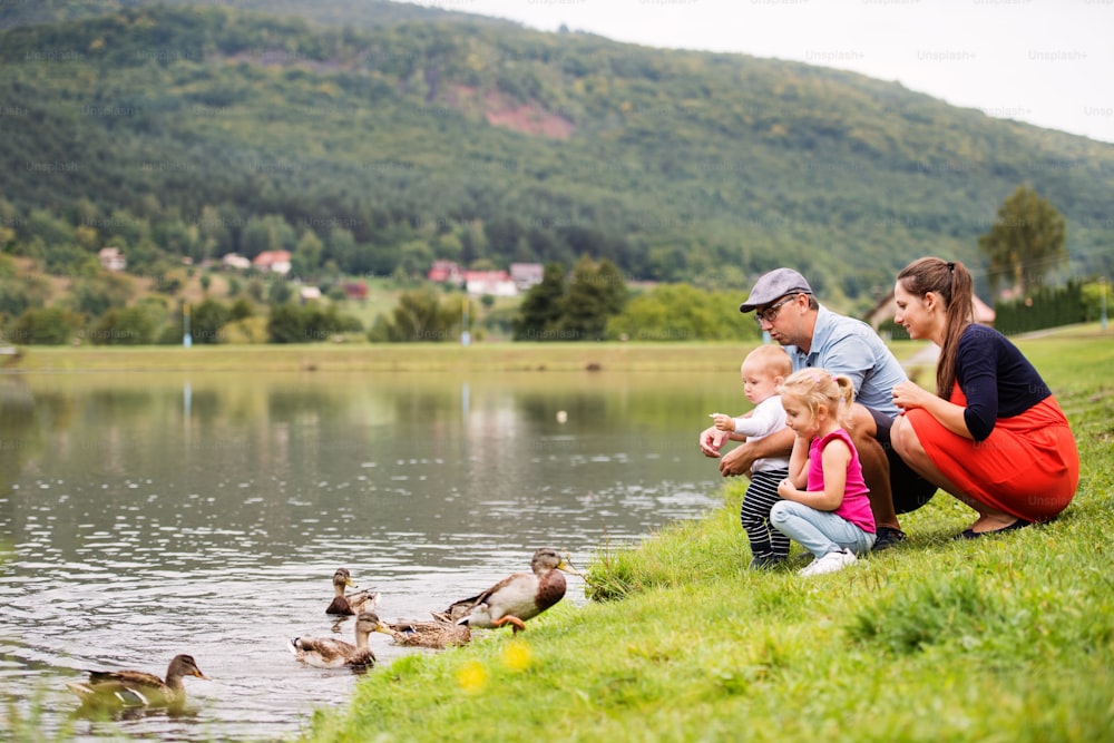 Happy young family spending time together outside in green nature, feeding ducks on a lake bank.