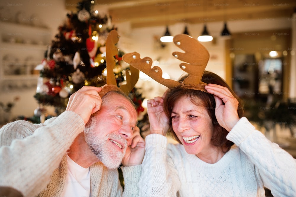 Un couple de personnes âgées assis par terre devant un arbre de Noël illuminé à l’intérieur de leur maison portant des bois de cerf, s’amusant.