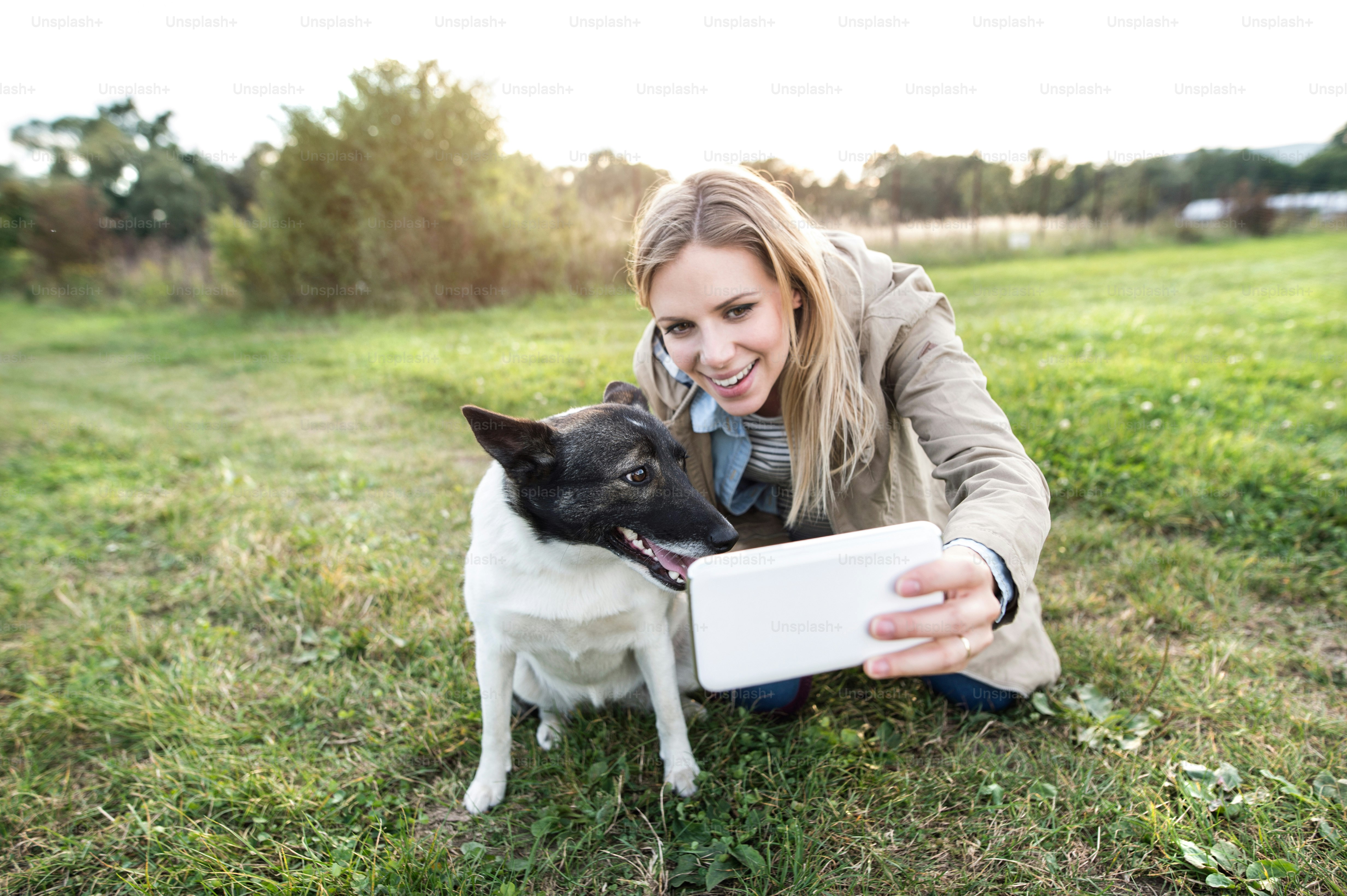 Beautiful young woman on a walk with a dog in green sunny nature, taking selfie with smart phone.