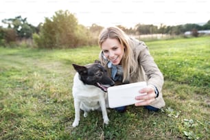 Belle jeune femme en promenade avec un chien dans une nature verte et ensoleillée, prenant selfie avec un téléphone intelligent.