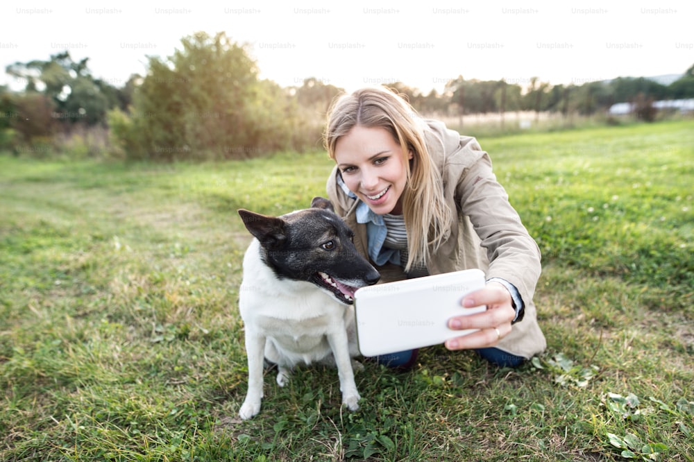 Beautiful young woman on a walk with a dog in green sunny nature, taking selfie with smart phone.