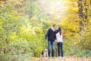 Beautiful young couple with dog on a walk in colorful sunny autumn forest, kissing.