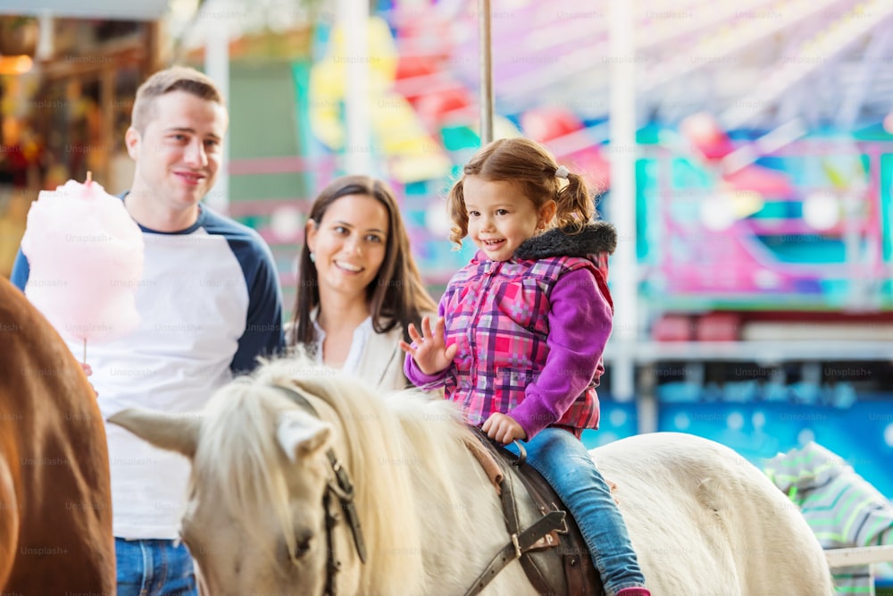 Little girl enjoying a ride on pony at fun fair, parents watching her, amusement park