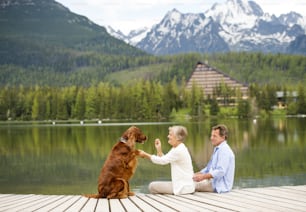 Pareja mayor con perro sentado en el muelle sobre el lago de montaña con montañas en el fondo