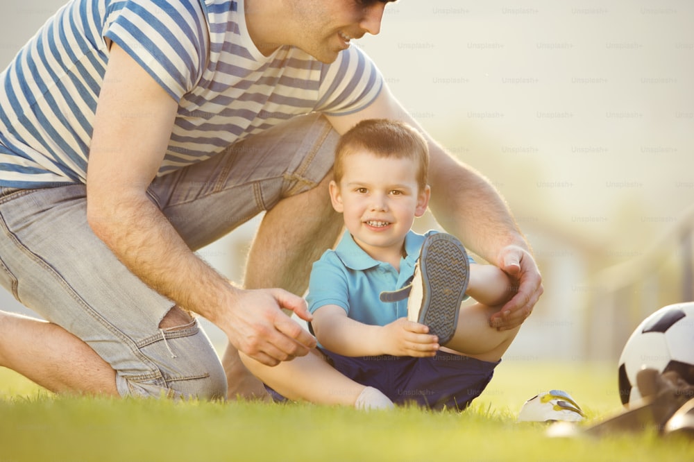 Padre joven con su hijo pequeño cambiando zapatos en el campo de fútbol