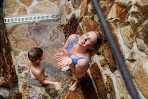 Draufsicht auf eine Frau mit seinem Sohn im Badeanzug, die an einem heißen Sommertag in der Außendusche im Garten duscht.