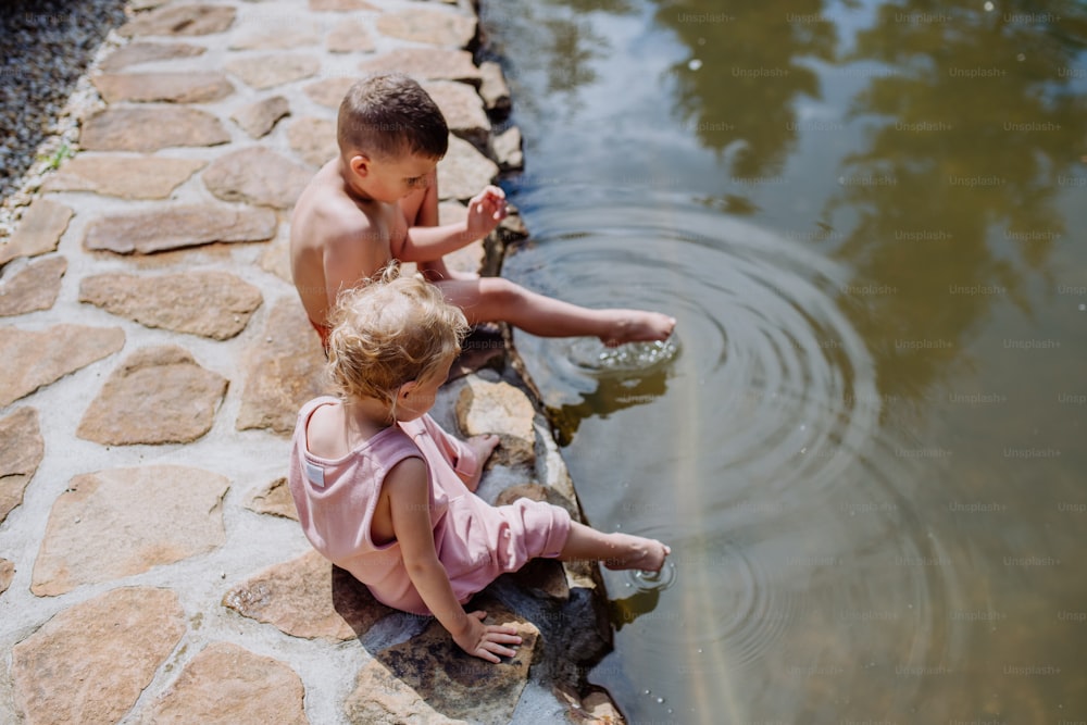 Ein süßes Mädchen und ein Junge sitzen zusammen auf dem Fußweg am See, lassen ihre Füße im Wasser baumeln, Sommerurlaubskonzept.