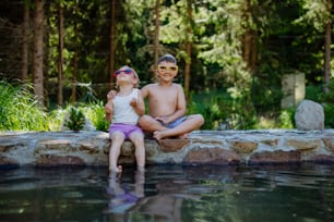 Ein süßes Mädchen und ein Junge sitzen zusammen auf dem Fußweg am See mit Sonnenbrille, lassen die Füße im Wasser baumeln, Sommerurlaubskonzept.