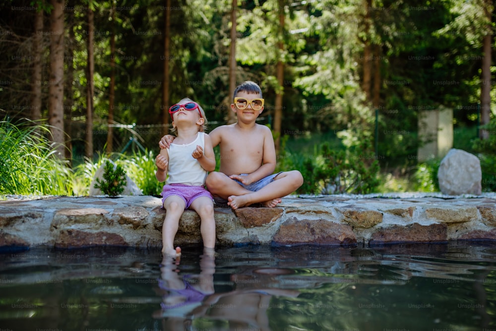 サングラスをかけた湖のほとりの歩道に一緒に座っているかわいい女の子と男の子が、水に足をぶら下げ、夏休みのコンセプト。