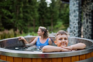 Mother with her little son enjoying bathing in a wooden barrel hot tub in the terrace of the cottage. Wooden bathtub with a fireplace to burn wood and heat water.