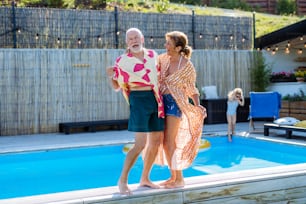 Um casal de idosos feliz se divertindo ao caminhar pela piscina durante as férias familiares da geração de verão.