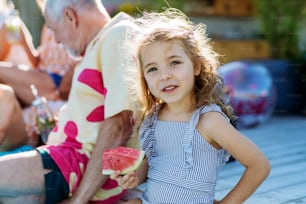 Cute little girl in a swimsuit eating watermelon corn and sitting next to pool with family at garden party.