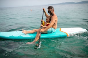 A happy father and little son on paddle board in sea together. Fatherhood and quality time with child concept.