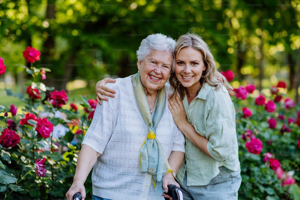 Ein Porträt einer erwachsenen Enkelin mit älterer Großmutter auf einem Spaziergang im Park, mit Rosen im Hintergrund