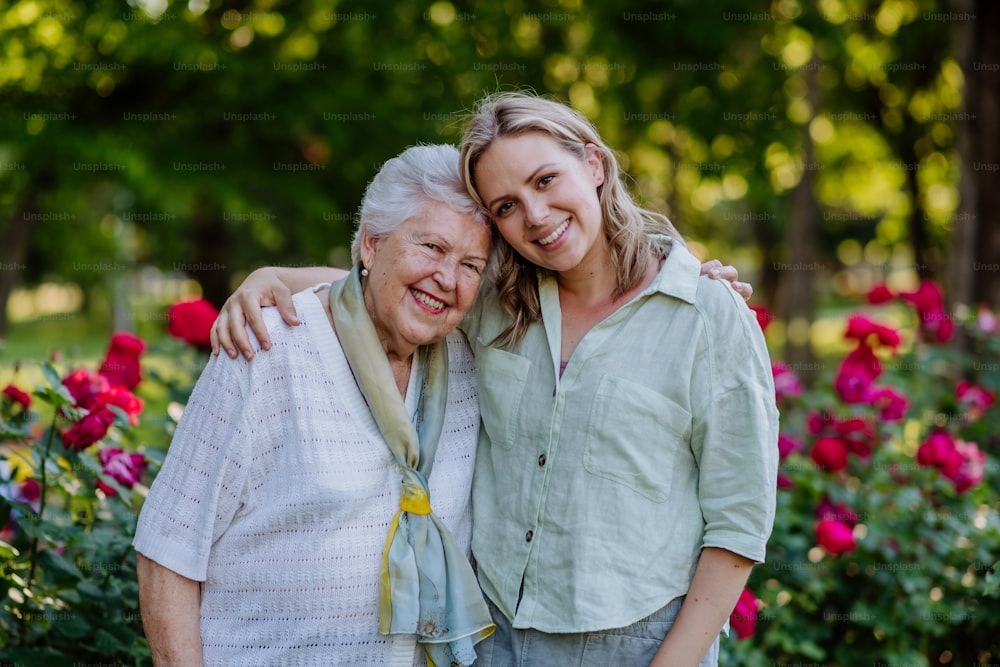 A portrait of adult granddaughter with senior grandmother on walk in park, with roses at background