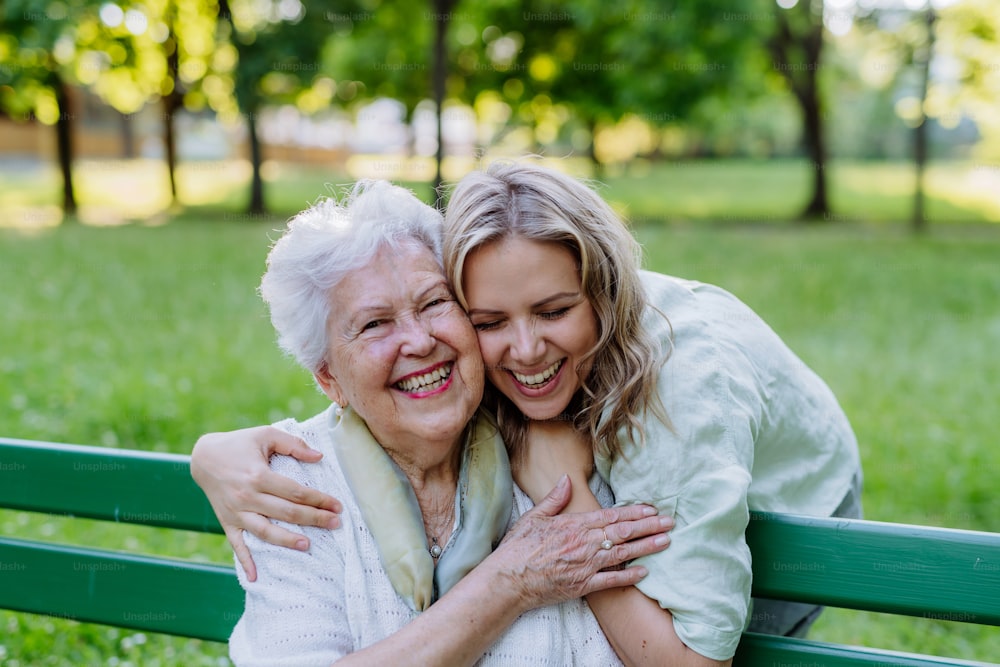 Ein Porträt einer erwachsenen Enkelin, die ihre ältere Großmutter umarmt, wenn sie auf einer Bank im Park sitzt.